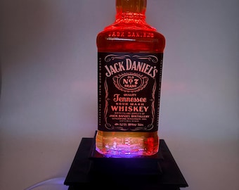 May include: A Jack Daniel's whiskey bottle illuminated with red and purple LED lights, resting on a black, tiered platform. The bottle's label is clearly visible, showcasing the brand's logo and product details. The overall effect is a striking display.