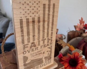 May include: Wooden plaque featuring an American flag design with a cross and the words "FAITH OVER FEAR" and "FREEDOM". Includes a portrait and the text "Rest in Power Charlie Kirk 1983-2023". The plaque is mounted on a wooden base.