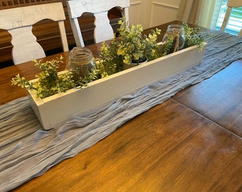 May include: A white wooden box planter filled with green plants and clear glass jars sits on a blue linen table runner on a wooden table.
