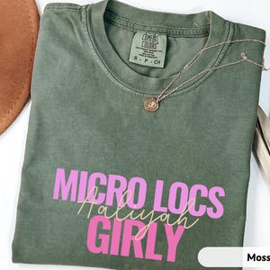 May include: A moss green t-shirt with the words "MICRO LOCS Haliyah GIRLY" in pink and gold lettering. A gold necklace with a pendant rests on the shirt. A brown hat and dried plant are in the background.