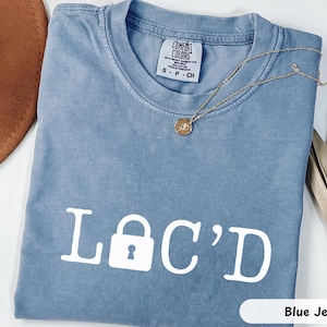 May include: A blue jean colored t-shirt with the word "LOCKED" printed in white, with a padlock symbol replacing the "O". A gold necklace with a pendant rests on the shirt. The shirt is a Comfort Colors brand.