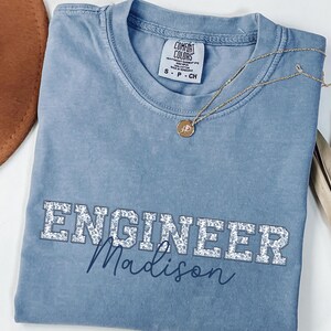 May include: A pale blue Comfort Colors t-shirt with the word "ENGINEER" in a floral design and the name "Madison" in cursive. A gold necklace with a pendant is on the shirt.