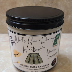 May include: A black candle jar with a white label that says "What's Your Damage Heather?" - Veronica. The label also features a plaid skirt, a hat, and a book. The candle is made with 100% soy wax and is hand-poured in New Jersey.