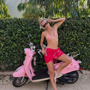 May include: A woman wearing a pink bikini top and a red and white patterned sarong sits on a pink scooter. She is wearing headphones and sunglasses. The scooter has a black seat and black wheels.