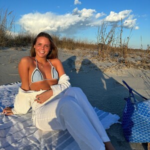 May include: A woman wearing a light blue crochet bikini top and white pants sits on a striped beach towel. She is on a sandy beach with tall grasses and a blue and white patterned tote bag beside her.