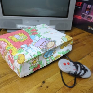 May include: A vintage Strawberry Shortcake themed video game console with a colorful floral print and a gray controller with red buttons.