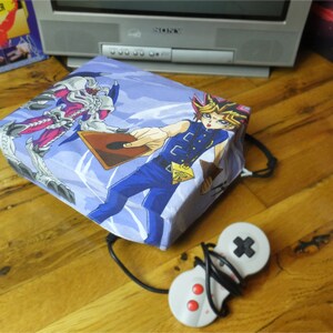 May include: A blue and purple fabric case with a graphic of a character from the Yu-Gi-Oh! anime series. The character is holding a card that says "The Dark Magician". The case is sitting on a wooden surface with a video game controller.