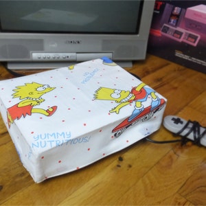 May include: A white gaming console with a Simpsons-themed cover featuring Bart and Lisa Simpson. The cover has red polka dots and the text "Yummy Nutritious!" The console is connected to a grey controller with a black cord.