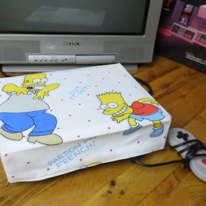 May include: White Nintendo Entertainment System (NES) console cover with a red, white, and blue Simpsons design. The cover features the characters Bart and Homer Simpson with the text "Cut it out!" and "Pardon my French!" printed on it. A black and gray NES controller is visible in the foreground.