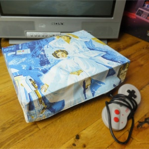 May include: A blue and white fabric cover with a Star Wars theme featuring Princess Leia and a Rebel pilot. The cover is designed to fit a gaming console.