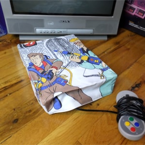 May include: A white and blue video game console cover with a cartoon image of the Ghostbusters characters. The cover is sitting on a wooden surface with a gray video game controller and a black cord.