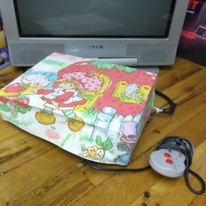 May include: A white and pink fabric gaming console cover with a strawberry shortcake design. The cover has a cord and a gray controller with two red buttons.