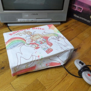 May include: A white fabric box with a rainbow and unicorn design. The box has the words "Rainbow Brite" printed on it. The box is sitting on a wooden surface.