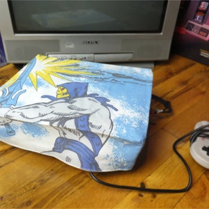 May include: A white fabric bag with a blue and yellow design featuring a cartoon character holding a sword. The character is wearing a blue cape and a helmet. The bag is on a wooden surface.