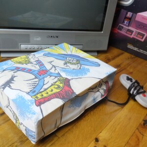 May include: A blue and white fabric cover with a graphic of He-Man, a fictional superhero, holding a sword. The cover is designed to fit a video game console.