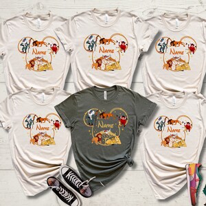 May include: Six t-shirts with a graphic of characters from The Lion King in a Mickey Mouse ear shape. The shirts are in various colors, including beige and green. The graphic includes the text "NAME" on each shirt.