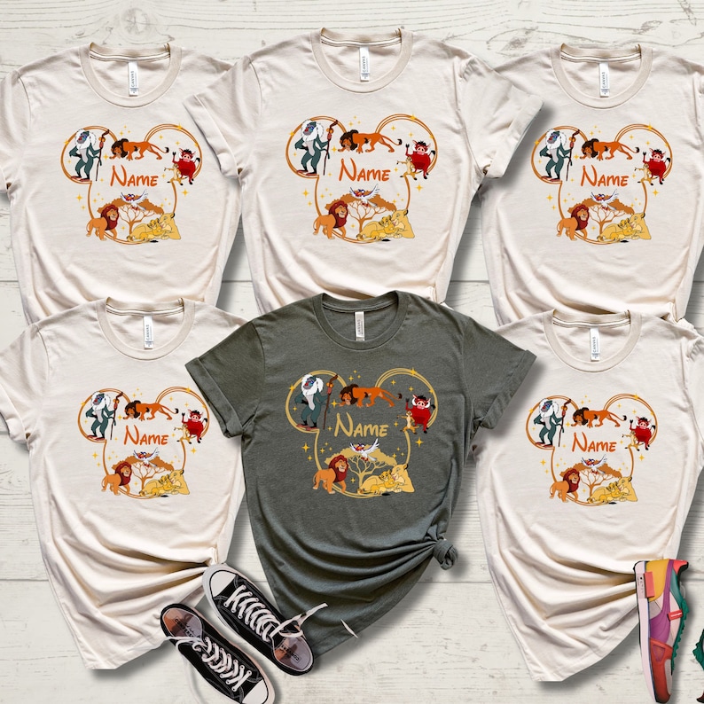 May include: Six t-shirts with a graphic of characters from The Lion King in a Mickey Mouse ear shape. The shirts are in various colors, including beige and green. The graphic includes the text "NAME" on each shirt.