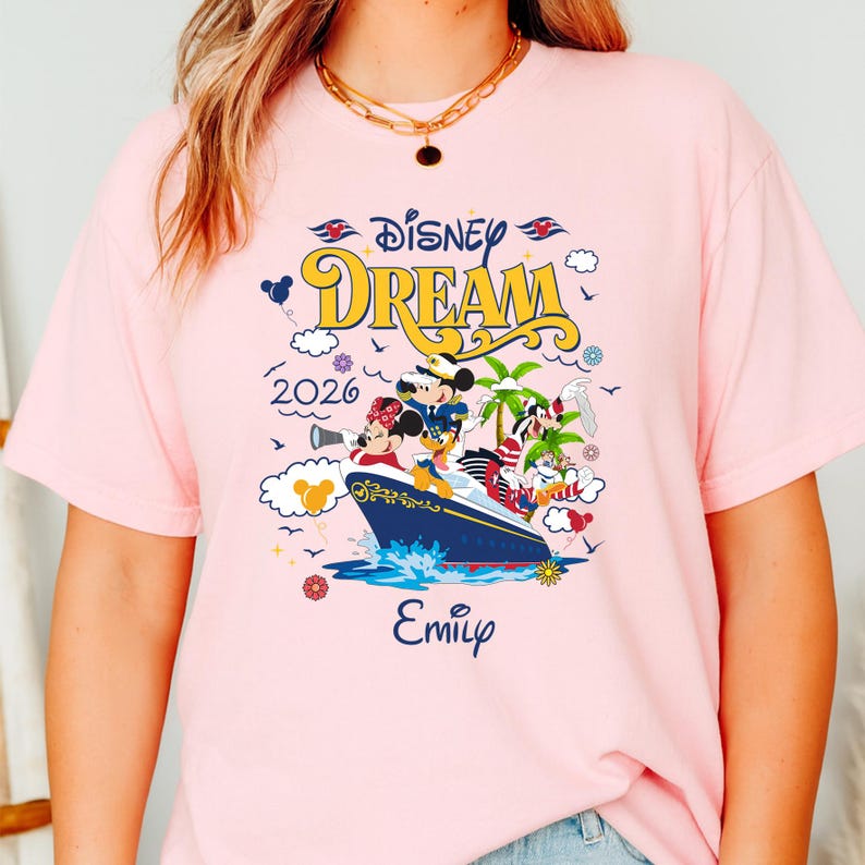 May include: Pink t-shirt featuring a colorful graphic of a cruise ship with Disney characters, the words "Disney Dream" and the year "2026". The name "Emily" is also printed on the shirt. The design includes Mickey Mouse, Minnie Mouse, and other characters.