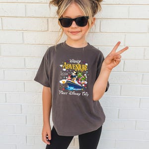 May include: A dark gray t-shirt featuring a colorful graphic of Disney characters on a boat, with the words "Disney Adventure 2026" and "First Disney Trip". The shirt is worn by a young person wearing sunglasses.