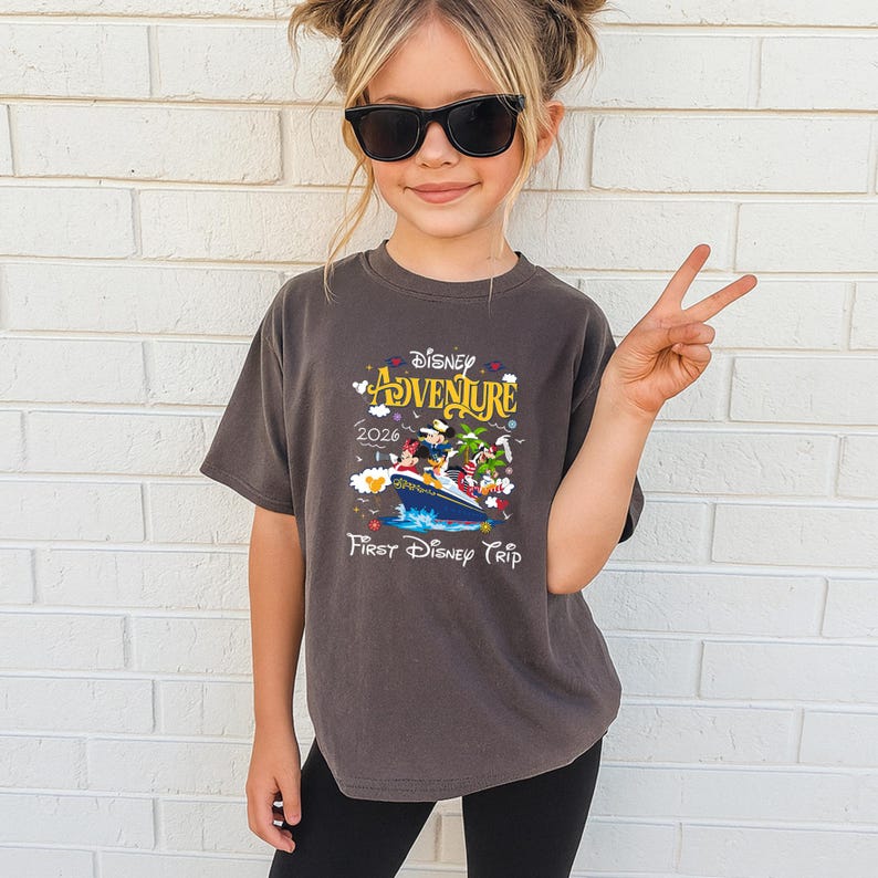 May include: A dark gray t-shirt featuring a colorful graphic of Disney characters on a boat, with the words "Disney Adventure 2026" and "First Disney Trip". The shirt is worn by a young person wearing sunglasses.