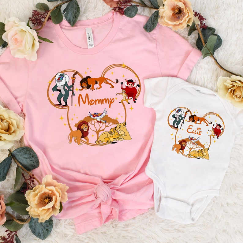 May include: Pink and white t-shirts with a Mickey Mouse ear design featuring characters from The Lion King. The pink shirt says "Mommy" and the white shirt says "Ellie".