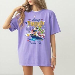 May include: A lilac-colored t-shirt featuring a Disney Fantasy cruise ship graphic. The graphic includes Mickey Mouse, Minnie Mouse, and other Disney characters. The text "Disney Fantasy 2026 Family Trip" is also printed on the shirt.
