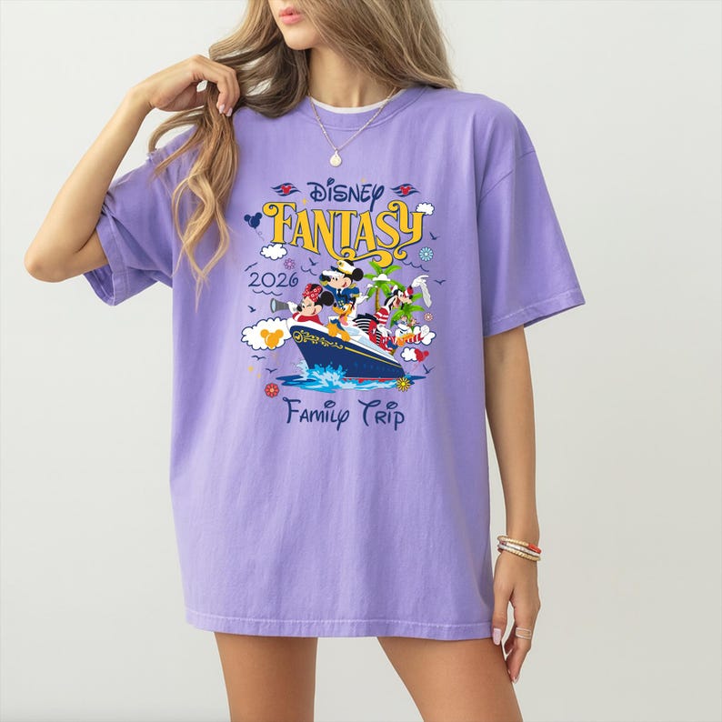 May include: A lilac-colored t-shirt featuring a Disney Fantasy cruise ship graphic. The graphic includes Mickey Mouse, Minnie Mouse, and other Disney characters. The text "Disney Fantasy 2026 Family Trip" is also printed on the shirt.