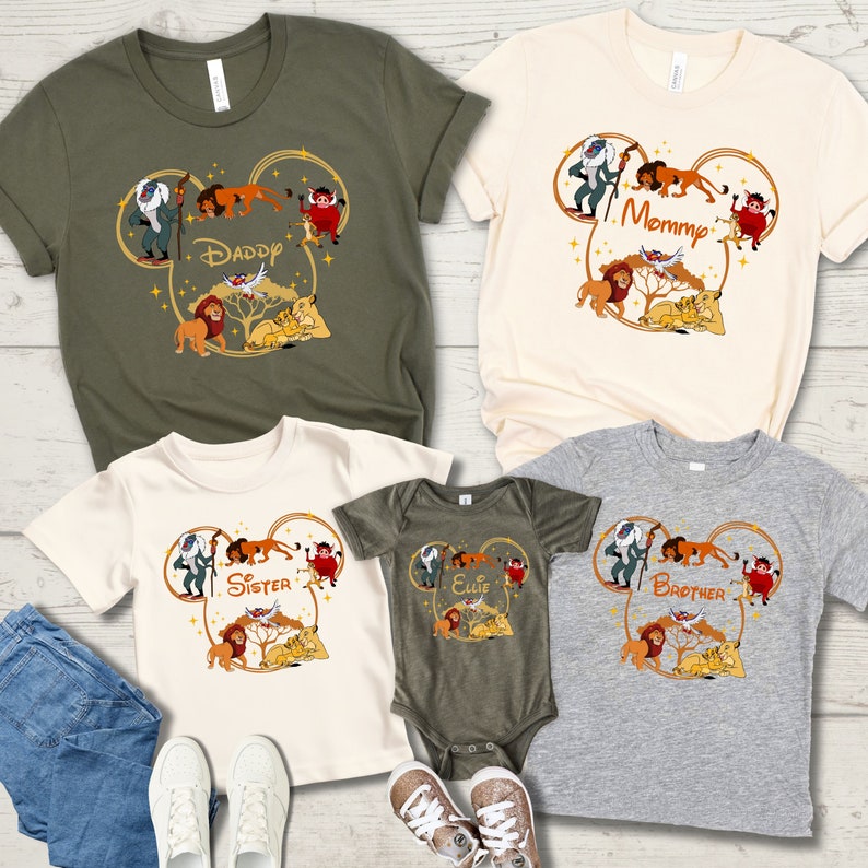 May include: Set of four t-shirts featuring a Mickey Mouse ear design with characters from The Lion King. The shirts are in green, beige, white, and gray with the words "Daddy", "Mommy", "Sister", and "Brother" printed on them.