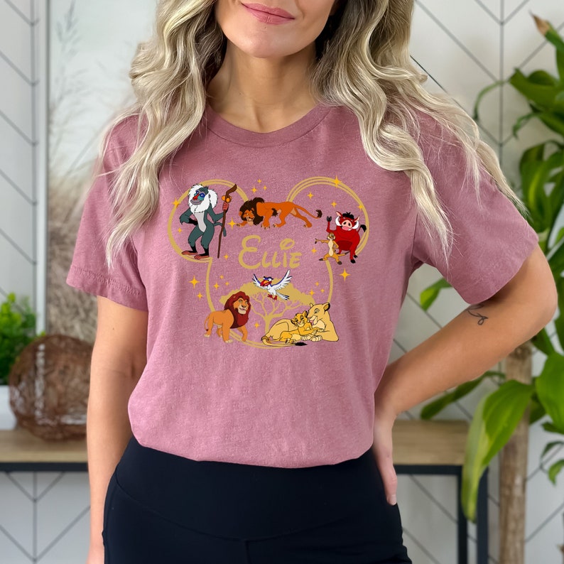 May include: A pink t-shirt with a gold Mickey Mouse head design featuring characters from The Lion King. The design includes Simba, Nala, Mufasa, Zazu, and Pumbaa. The text "Ellie" is written inside the Mickey Mouse head.