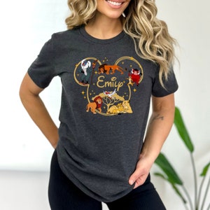 May include: A dark grey t-shirt with a gold Mickey Mouse ear silhouette featuring characters from The Lion King, including Simba, Mufasa, Pumbaa, and Timon. The name "Emily" is printed inside the silhouette.