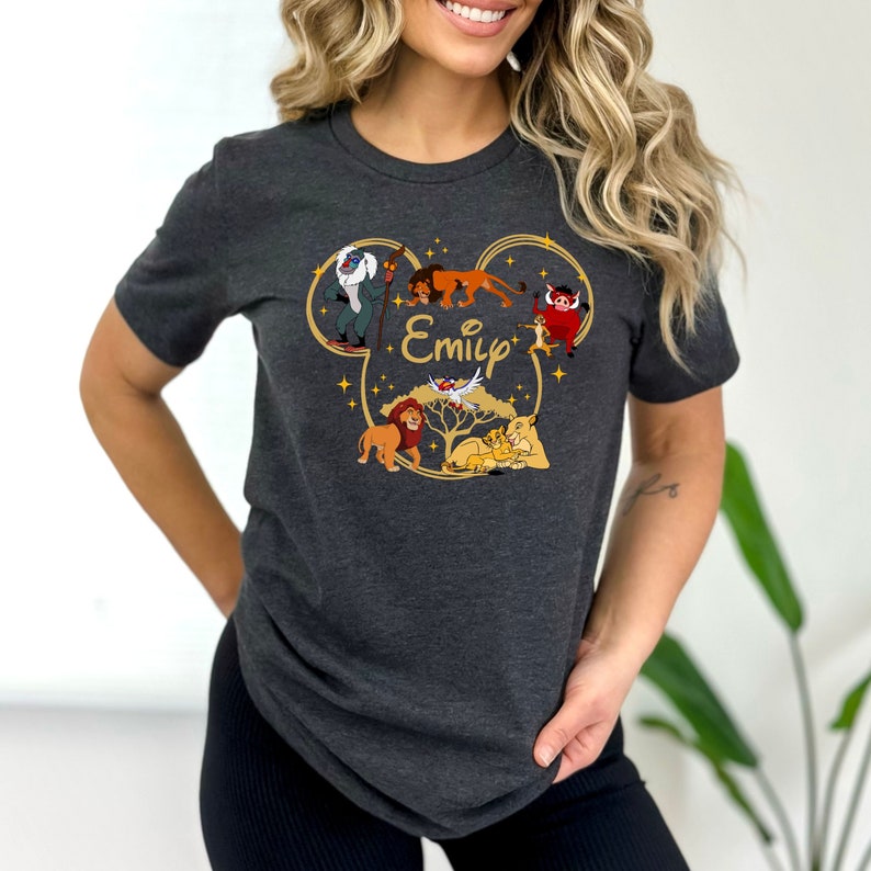 May include: A dark grey t-shirt with a gold Mickey Mouse ear silhouette featuring characters from The Lion King, including Simba, Mufasa, Pumbaa, and Timon. The name "Emily" is printed inside the silhouette.