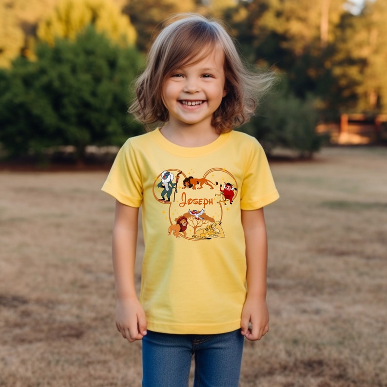 May include: Yellow short-sleeved t-shirt with a Mickey Mouse head design featuring characters from The Lion King. The design includes Simba, Mufasa, Pumbaa, Timon, and Rafiki. The name "Joseph" is printed on the shirt.