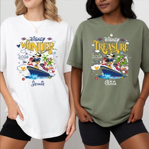 May include: Two t-shirts, one white and one olive green, featuring a Disney cruise design. The white shirt says "Disney Wonder 2026" with a ship graphic and the name "Jennie". The green shirt says "Disney Treasure 2026" with a ship graphic and the name "Olivia".
