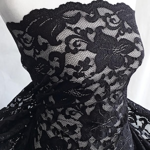 May include: Black lace fabric with a floral pattern. The delicate lace is draped over a mannequin, showcasing its intricate design and texture. The fabric is a deep black color, with a scalloped edge.