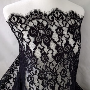 May include: Black lace fabric with a floral pattern. The lace is draped over a white mannequin, showcasing the intricate design and delicate texture. The fabric's edge has a scalloped trim.