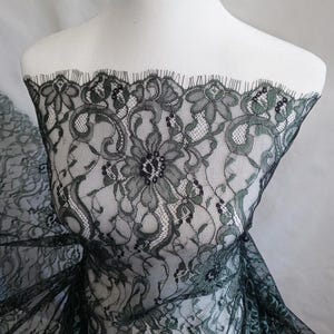 Puede incluir: Tela de encaje floral verde oscuro con borde festoneado. El encaje presenta un patrón detallado de flores y diseños en espiral. La tela está drapeada sobre un maniquí blanco, mostrando su textura y diseño. El encaje es semitransparente.