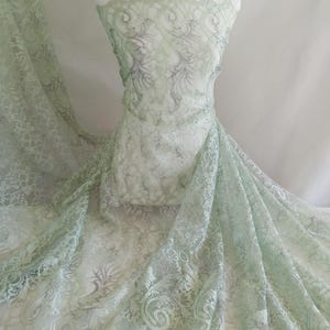 Puede incluir: Tela de encaje verde pálido con un intrincado diseño floral, drapeada sobre un maniquí. El encaje tiene un borde festoneado y es semitransparente. El tejido es elegante y ligero, ideal para confección o decoración.