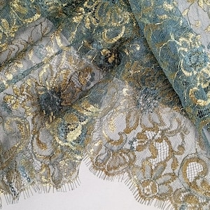 Può includere: Primo piano di un tessuto di pizzo verde acqua e oro. Il delicato pizzo presenta intricati motivi floreali con accenti dorati metallici. Il tessuto è piegato, rivelando la trama e il design dettagliati.
