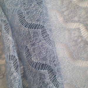 May include: Close-up of a light blue lace fabric. The delicate lace features an intricate floral and geometric pattern. The fabric is slightly translucent, revealing a lighter background in some areas. The texture appears soft and flowing.