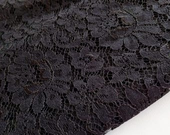 Puede incluir: Primer plano de tela de encaje negro. El encaje presenta un patrón floral con diseños detallados y delicados. La tela es de color negro intenso, con una superficie ligeramente texturizada. La imagen está bien iluminada, destacando los intrincados detalles del encaje.