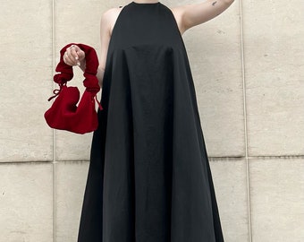 May include: A black halter-neck maxi dress with a flared skirt. A red handbag is held in the left hand. The person is wearing black sandals. The background is a light beige wall.