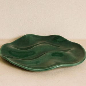 May include: A dark green ceramic dish with a wavy, organic shape. The dish has a glossy finish and is designed with flowing curves. This decorative piece is suitable for serving or display.