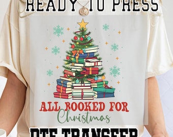 May include: A cream-colored t-shirt with a design of a Christmas tree made of books. The text "Ready to Press" is at the top of the shirt. The text "All Booked for Christmas" is below the tree. The text "DTG Transfer" is at the bottom of the shirt.