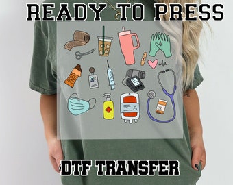 May include: Green t-shirt with a DTF transfer design featuring medical-themed illustrations. The design includes a bandage, scissors, a coffee cup, a pink tumbler, gloves, a heart, a stethoscope, and other medical supplies. The words "READY TO PRESS" and "DTF TRANSFER" are printed on the shirt.