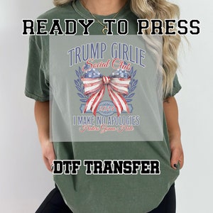 May include: A green t-shirt with a DTF transfer design featuring a red, white, and blue bow with a wreath and the text "Trump Girlie Social Club 2024 I Make No Apologies Haters Gonna Hate".