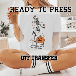 May include: White t-shirt with a DTF transfer design featuring a skeleton dancing on top of a pumpkin with the text "It's the most wonderful time of the year".