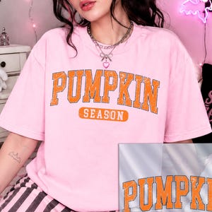 K&ouml;nnte beinhalten: Ein hellrosa Kurzarm-T-Shirt mit dem Text PUMPKIN SEASON in einer abgenutzten orangefarbenen College-Schriftart. Das Design ist mittig auf der Brust platziert. Eine separate Grafik&uuml;berlagerung zeigt dasselbe orangefarbene Textdesign.