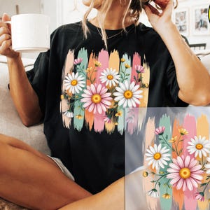 May include: Black t-shirt featuring a colorful floral design with daisies and brushstroke accents. The design includes pink, yellow, and white flowers with green stems and leaves. The image also shows a DTF transfer.