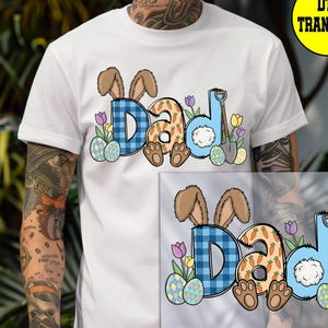 May include: White t-shirt with a colorful Easter-themed graphic. The design spells "DAD" with bunny ears, carrot-decorated letters, and a fluffy tail. Easter eggs, flowers, and a shovel complete the festive look.