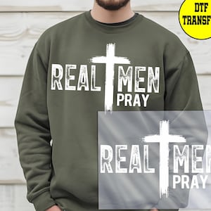 May include: Olive green sweatshirt with the text "REAL MEN PRAY" in white, featuring a white cross. The design is a DTF transfer. The sweatshirt is a casual garment.